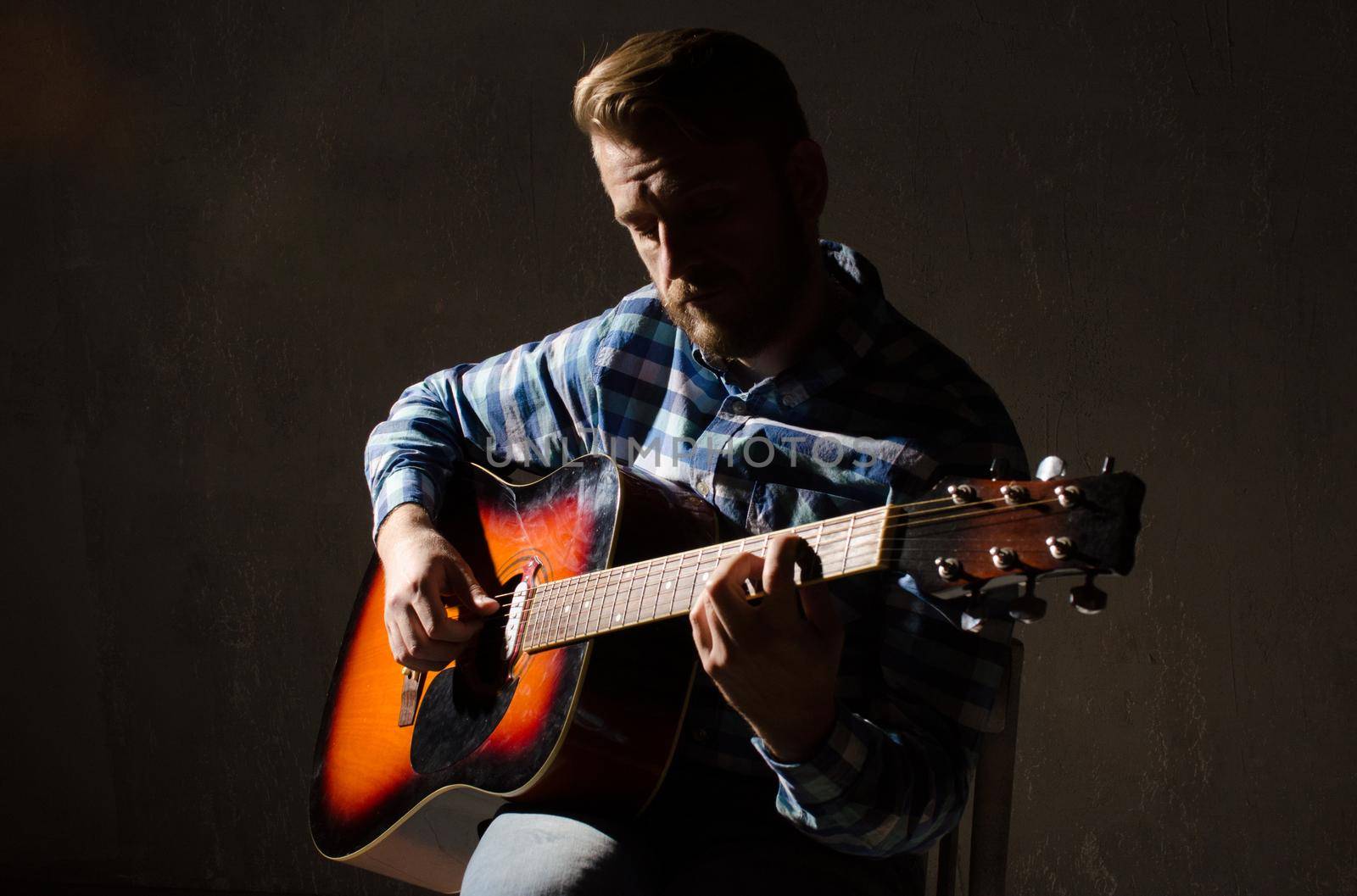 Adult man in a plaid shirt playing acoustic guitar. Low key portrait.