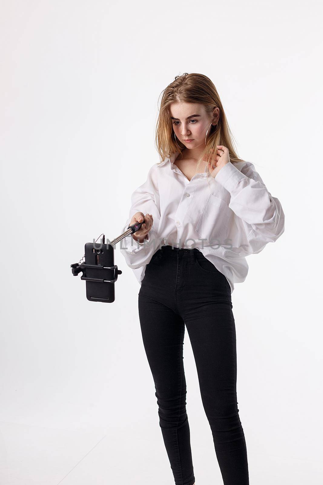 cute girl in shirt, jeans makes self portrait with smartphone attached to selfie stick. young pretty blond female with long hair poses using cell phone isolated on white background. teen communication