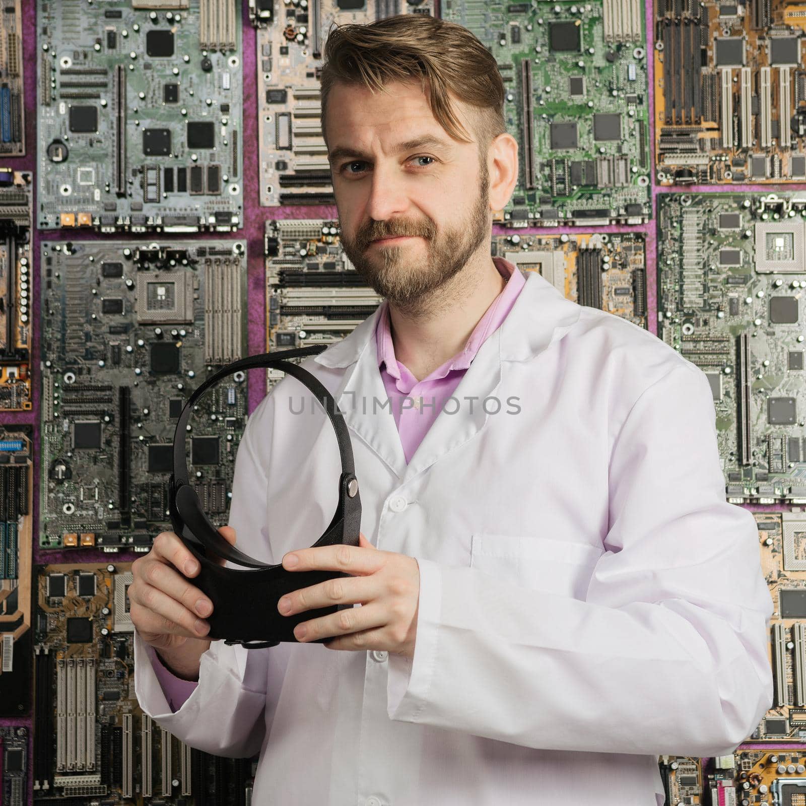 A young electronics engineer stands with a magnifying glass in his hands near the wall of motherboards.