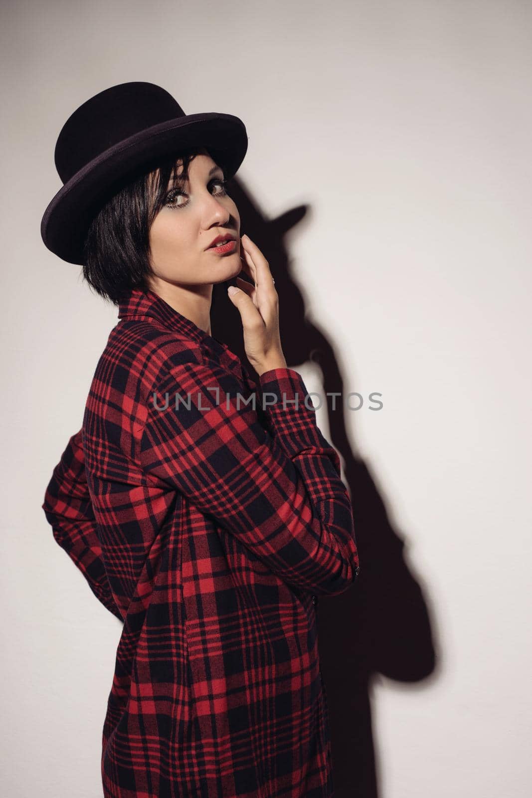 Attractive girl in red checkered shirt and hat bowler stands near white wall raising her hand to face