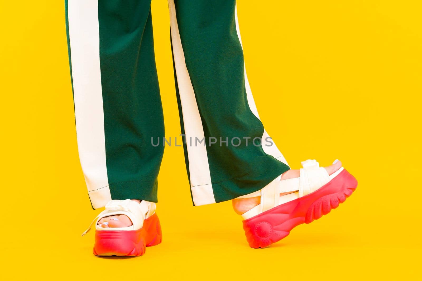 Women's legs in sport sandals with red soles and green pants with stripes on a yellow background.