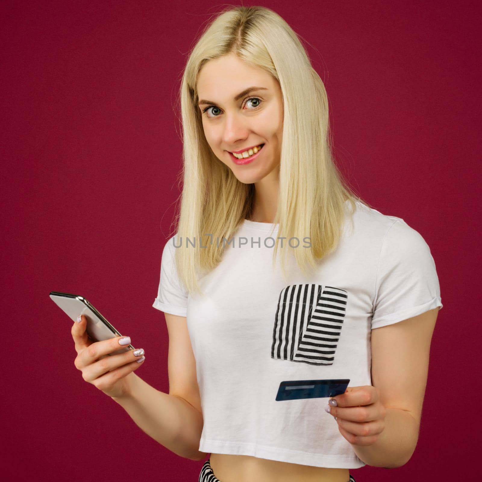 Young smiling woman buys online. Holds a smartphone and a credit card in hand on ruby background
