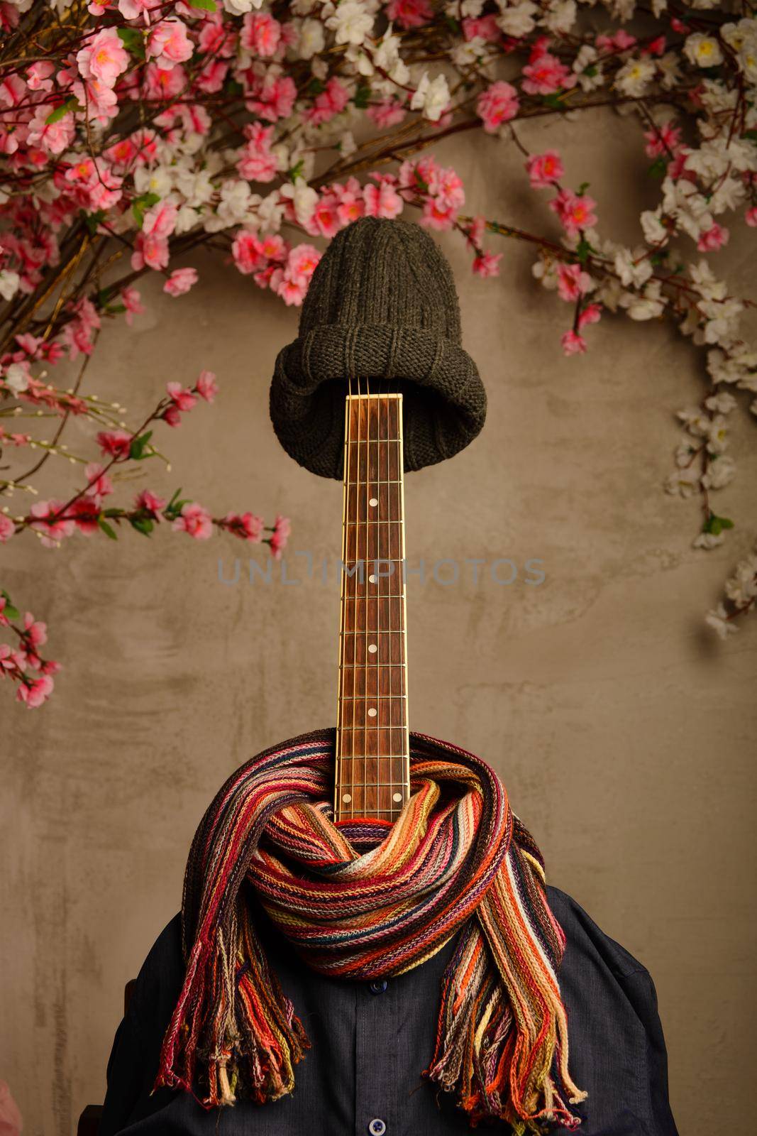 guitar dressed in a man's blue shirt with a scarf and a knitted cap next to a wall of flowers.