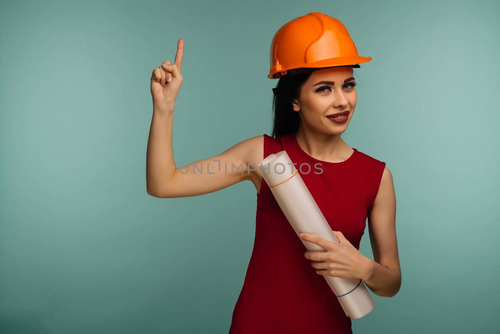 Young female engineer in orange helmet with drawings points finger up isolated on blue background - Image