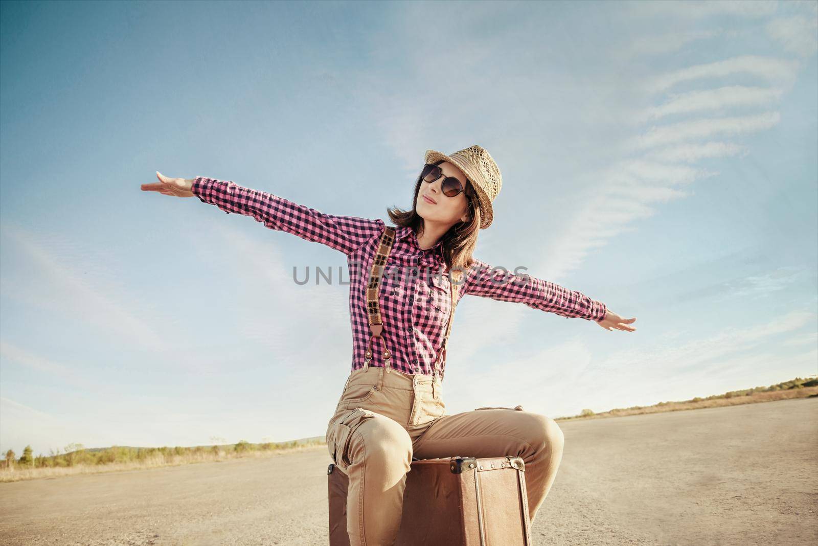 Happy hipster girl sits on vintage suitcase on road and makes a gesture of flight