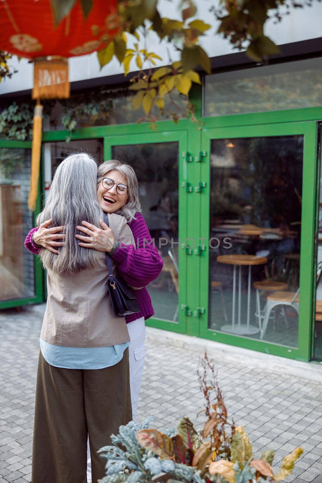 Joyful senior lady in warm jacket hugs long haired best friend meeting on modern city street. Long-time friendship relationship