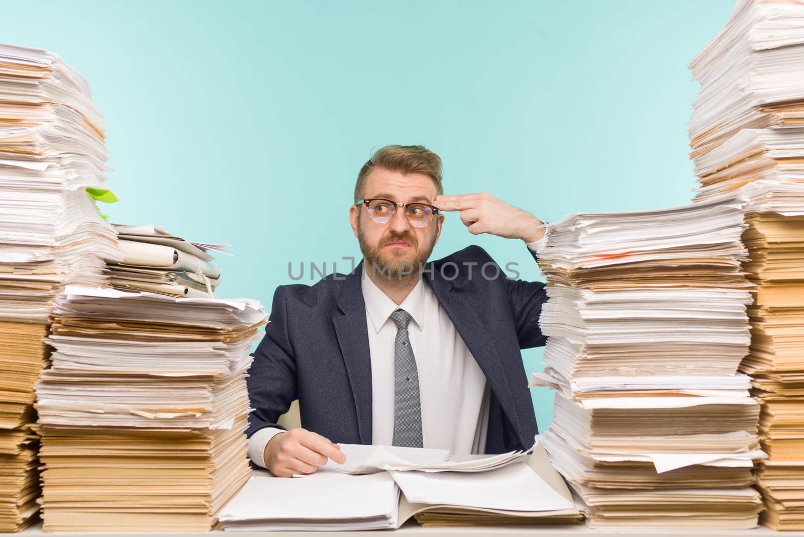 Shocked businessman sitting at the table with many papers in office, he is overloaded with work - image