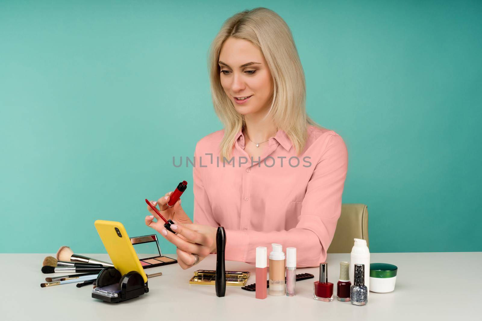 Candid shot of cute young Caucasian woman blogger presenting beauty products and broadcasting live video to social network, using lip gloss while recording everyday make up tutorial