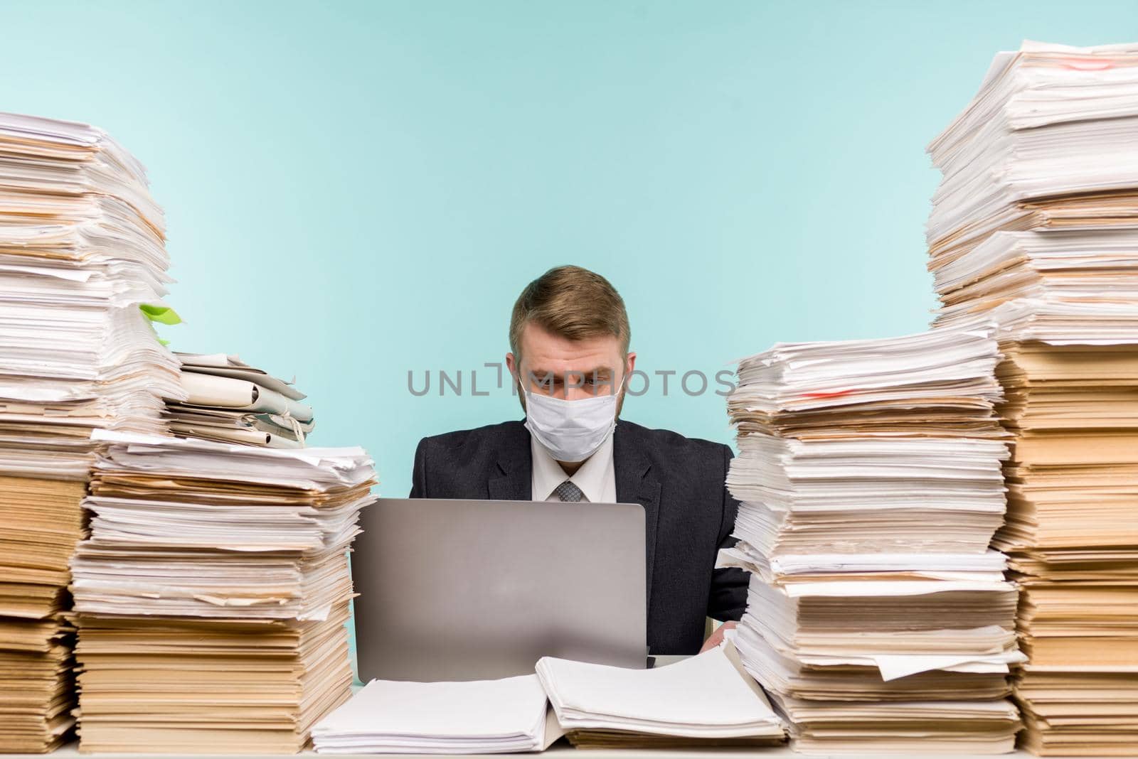 A male accountant or company manager works in an office in a pandemic in view of the accumulated paper work. A protective medical mask is on the face. On the desktop are large stacks of documents. - image