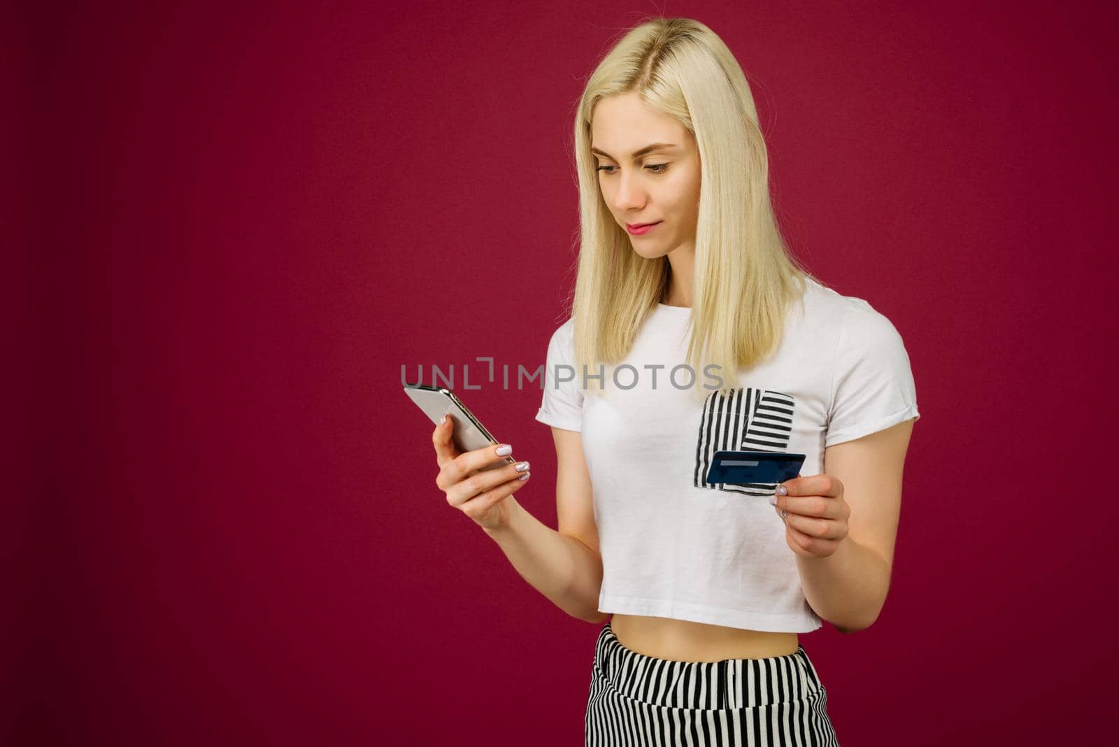 Young smiling woman buys online. Holds a smartphone and a credit card in hand on ruby background