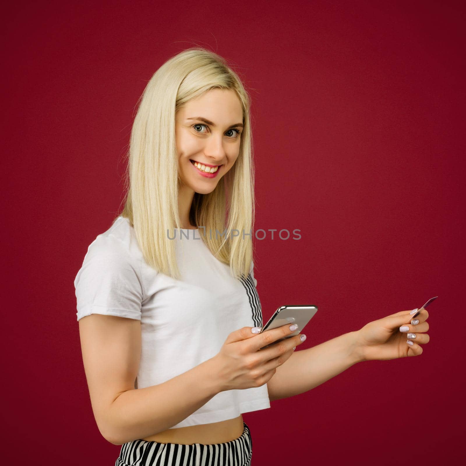 Young smiling woman buys online. Holds a smartphone and a credit card in hand on ruby background