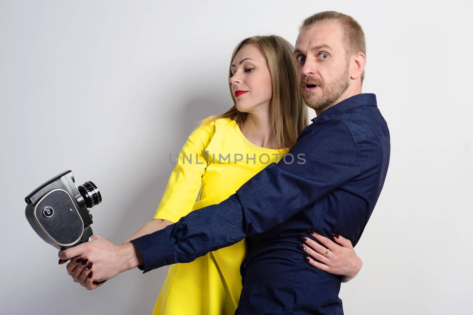 Surprised man in a blue shirt and a young slim girl in a yellow dress are hugging and shooting themselves on an old movie camera