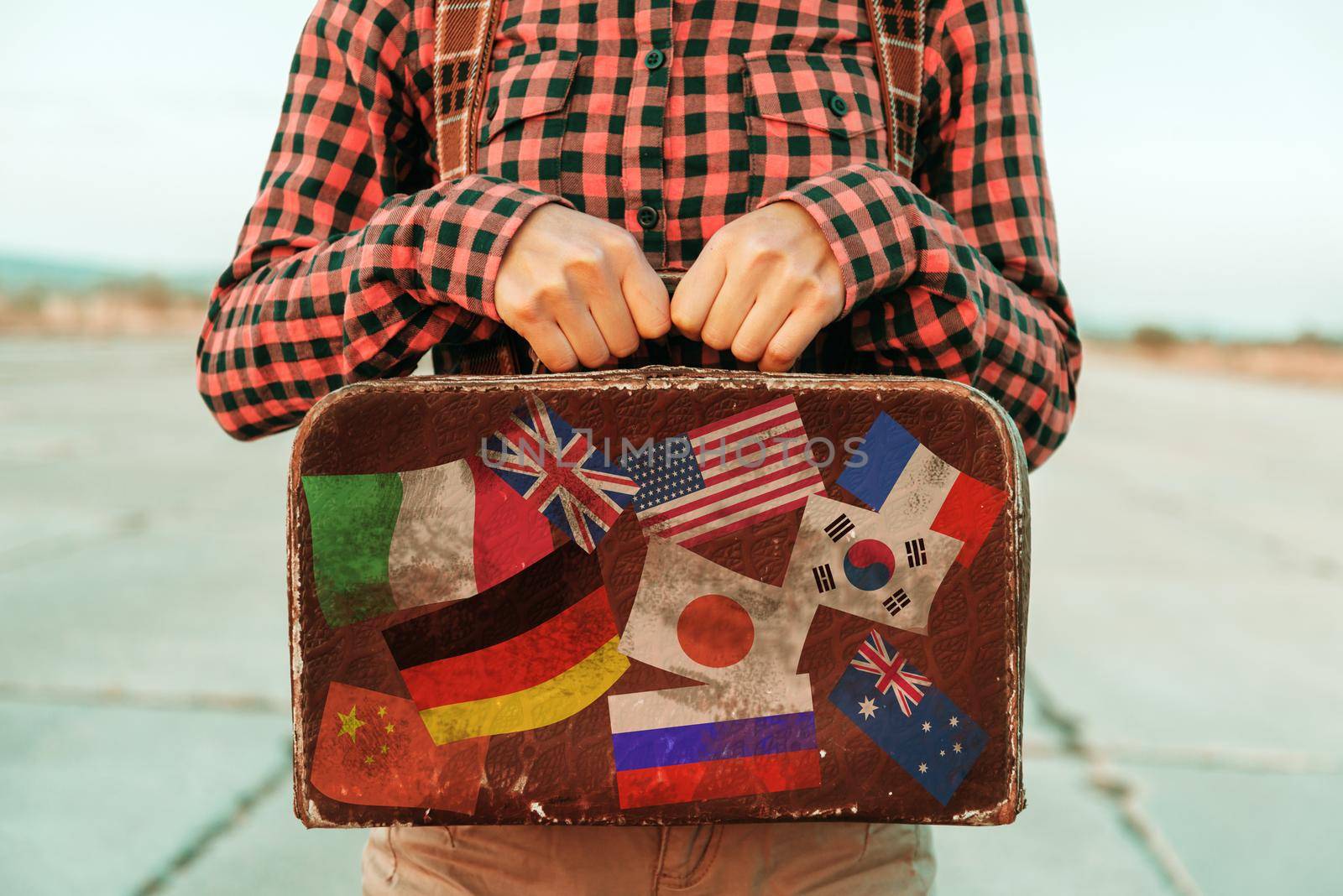 Woman holds small retro suitcase on road, face is not visible. Suitcase with stamps flags representing each country traveled.