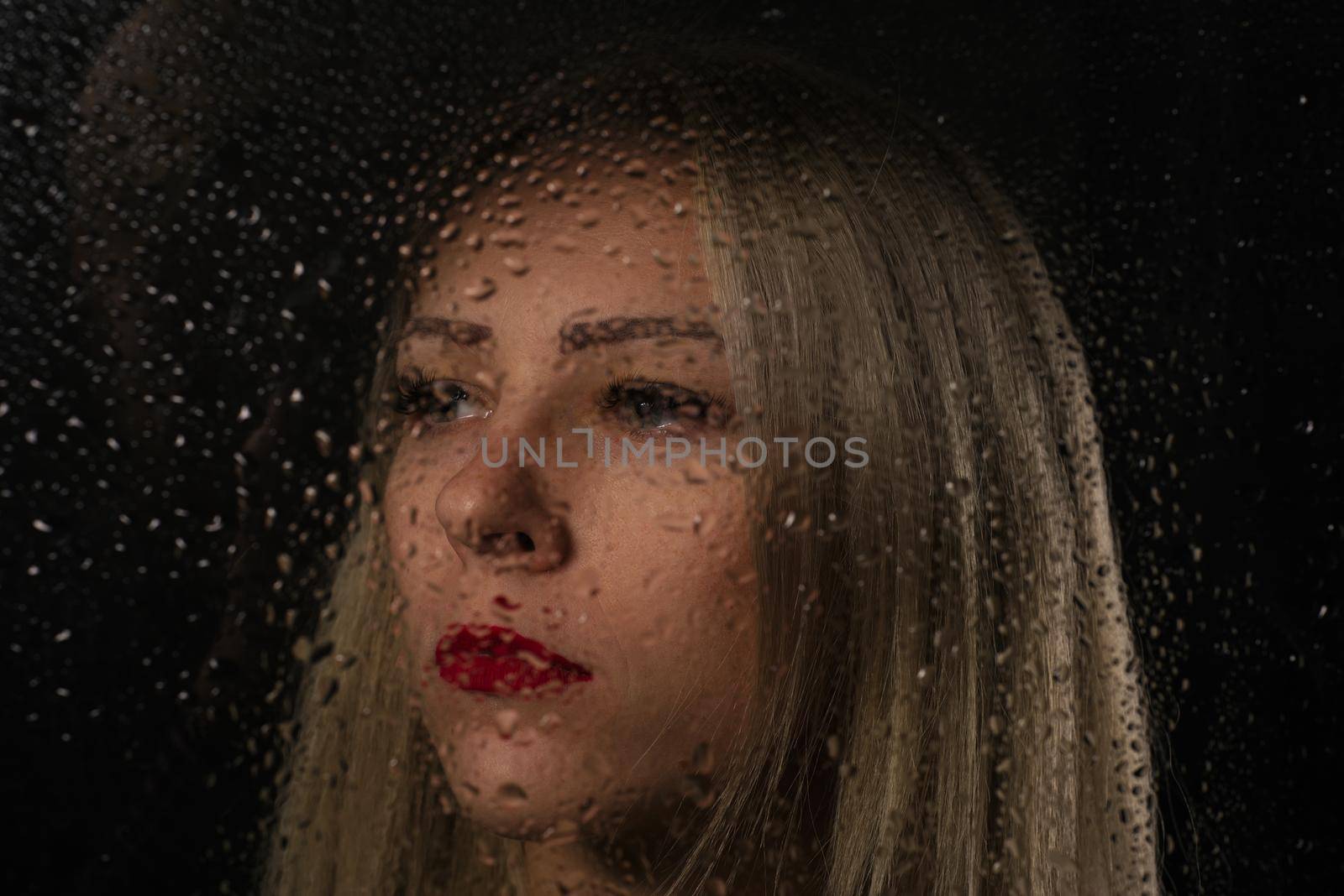 Smooth portrait of crying girl behind transparent glass covered by water drops.