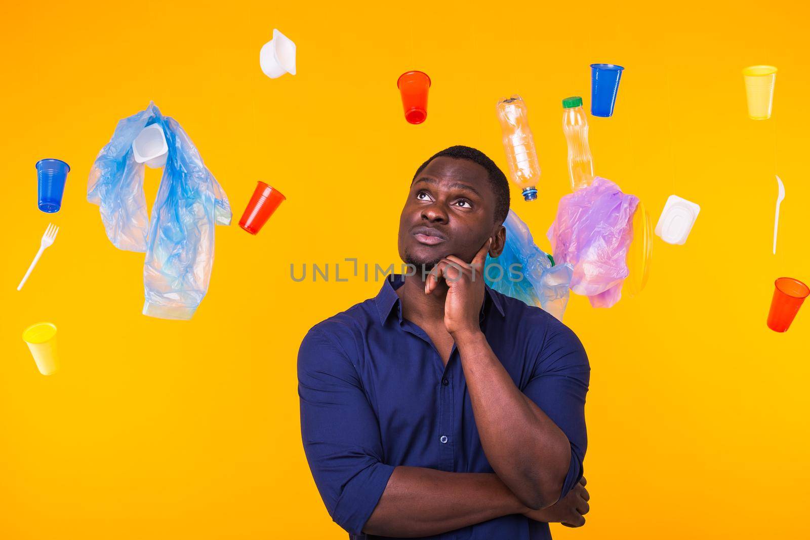 Problem of trash, plastic recycling, pollution and environmental concept - Serious african american man looking on trash on yellow background. He is thinking about ecology.