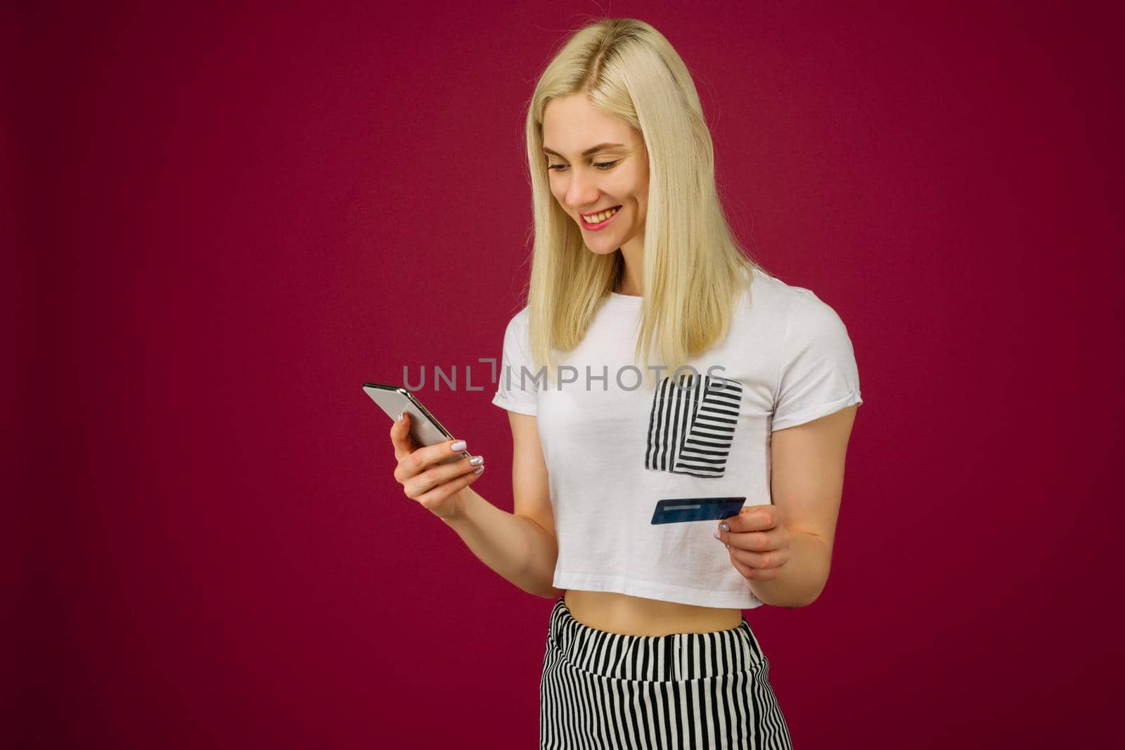 Young smiling woman buys online. Holds a smartphone and a credit card in hand on ruby background