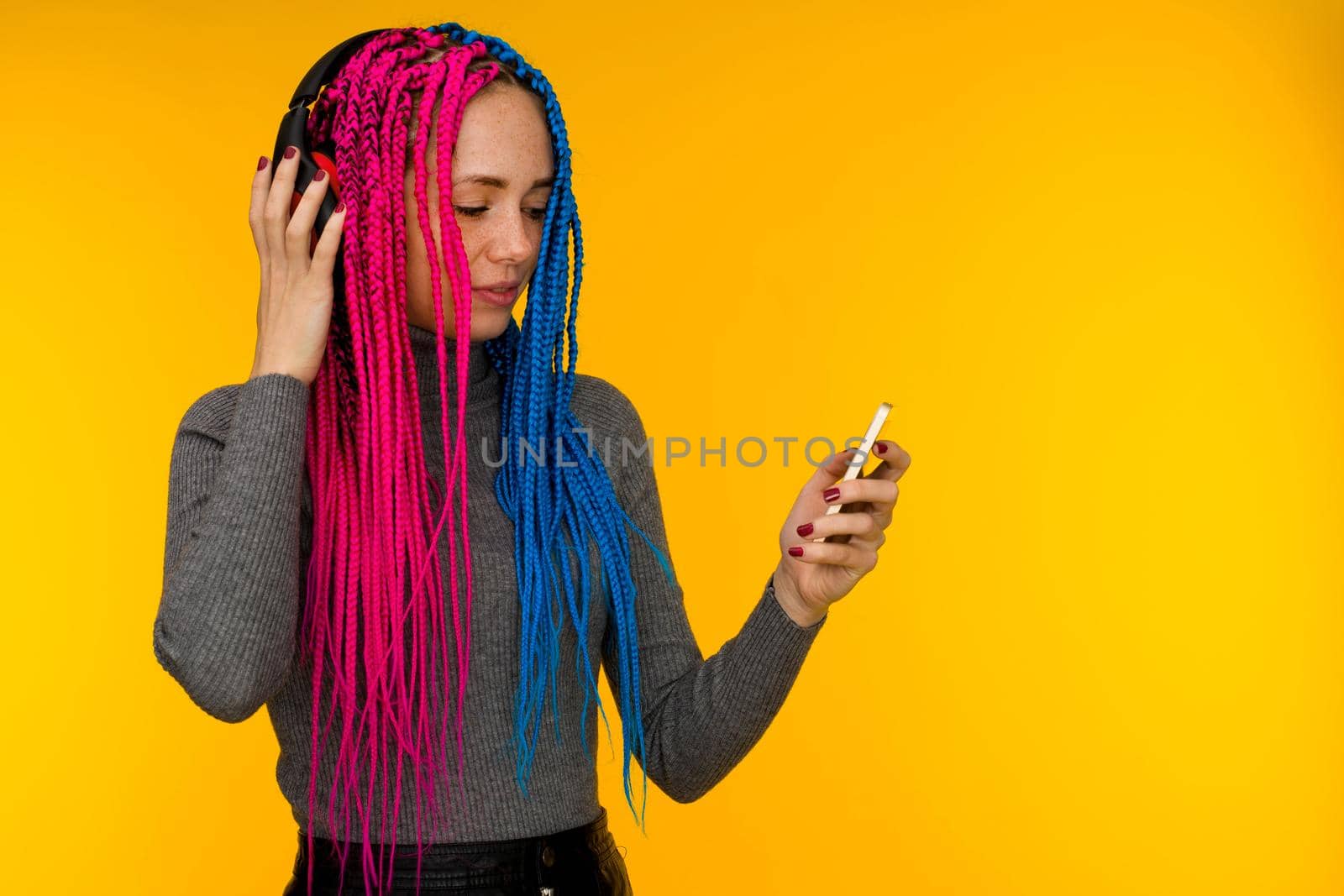 Happy cheerful woman with senegalese braids and freckles wearing wireless headphones listening to music from smartphone studio shot isolated on yellow background