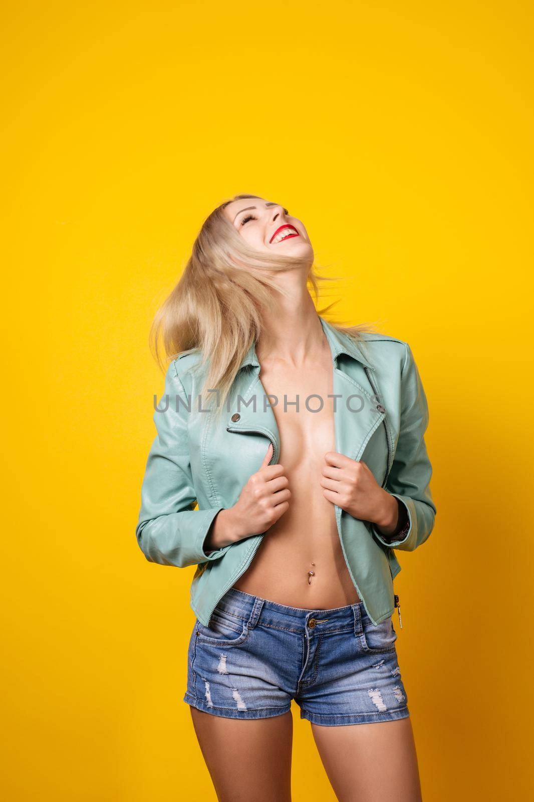Sexy young topless woman in leather jacket and short denim shorts posing on a yellow background