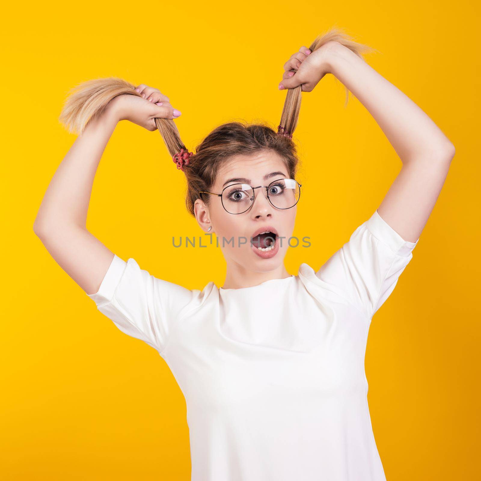 A young girl in glasses with her mouth open open in surprise holds her tails isolated on a yellow background