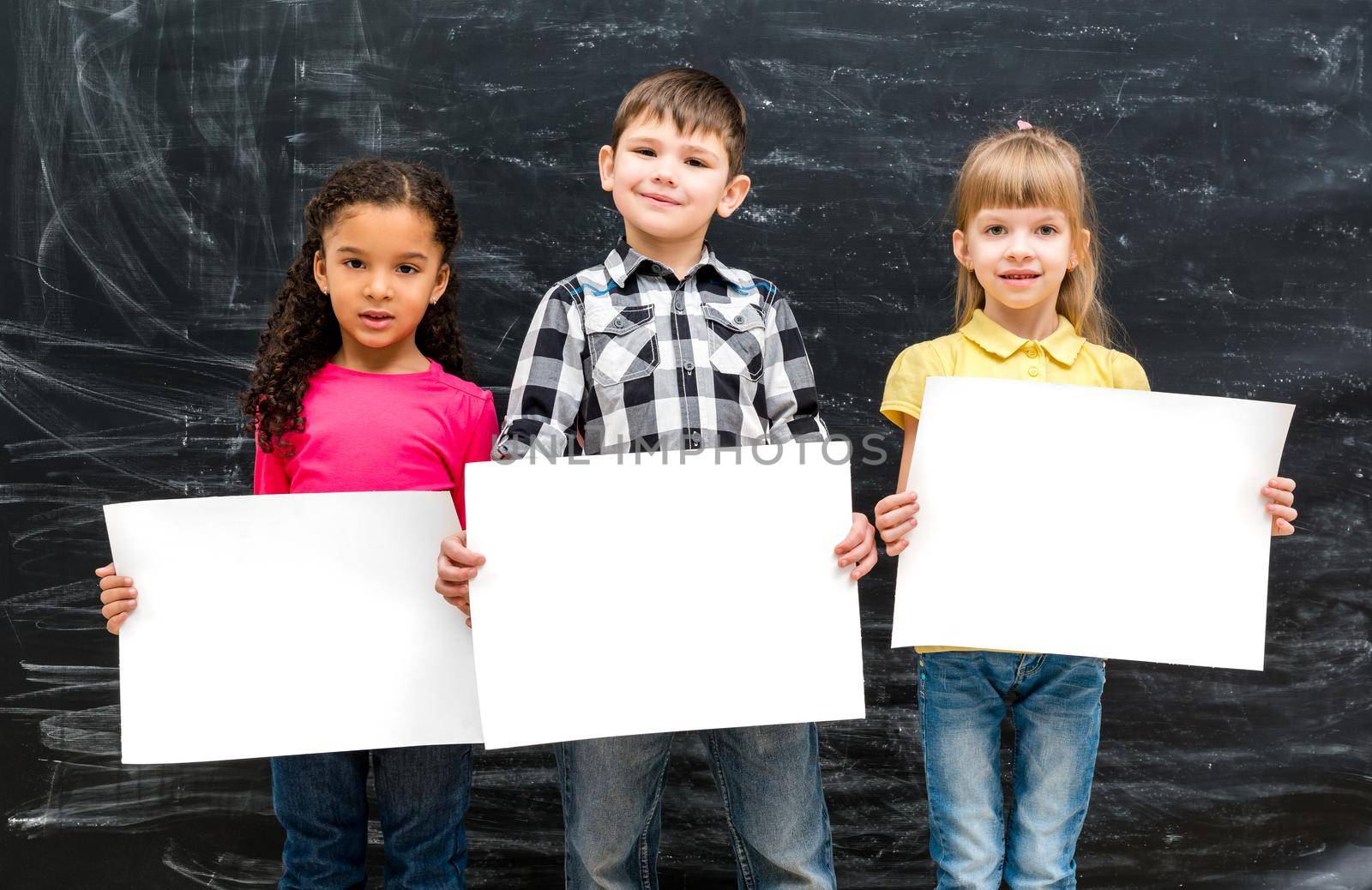 three cute children with empty paper sheets in hands and chalky blackboard on background