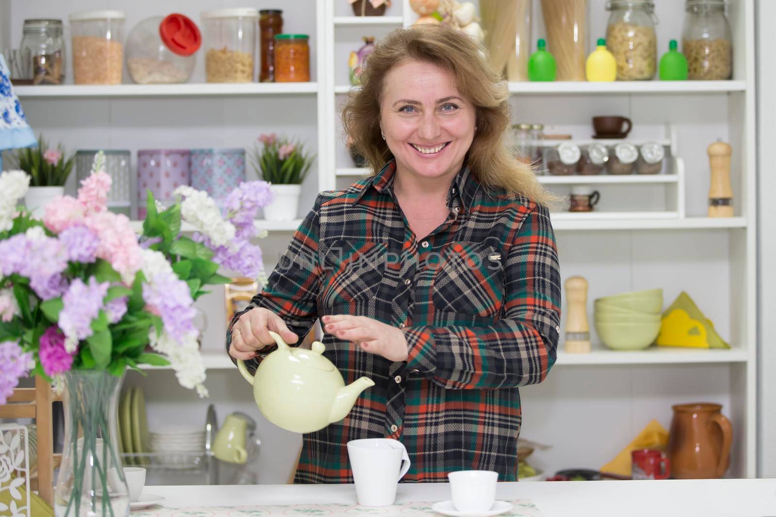 Beautiful happy ordinary elderly woman in the kitchen with a kettle and a cup