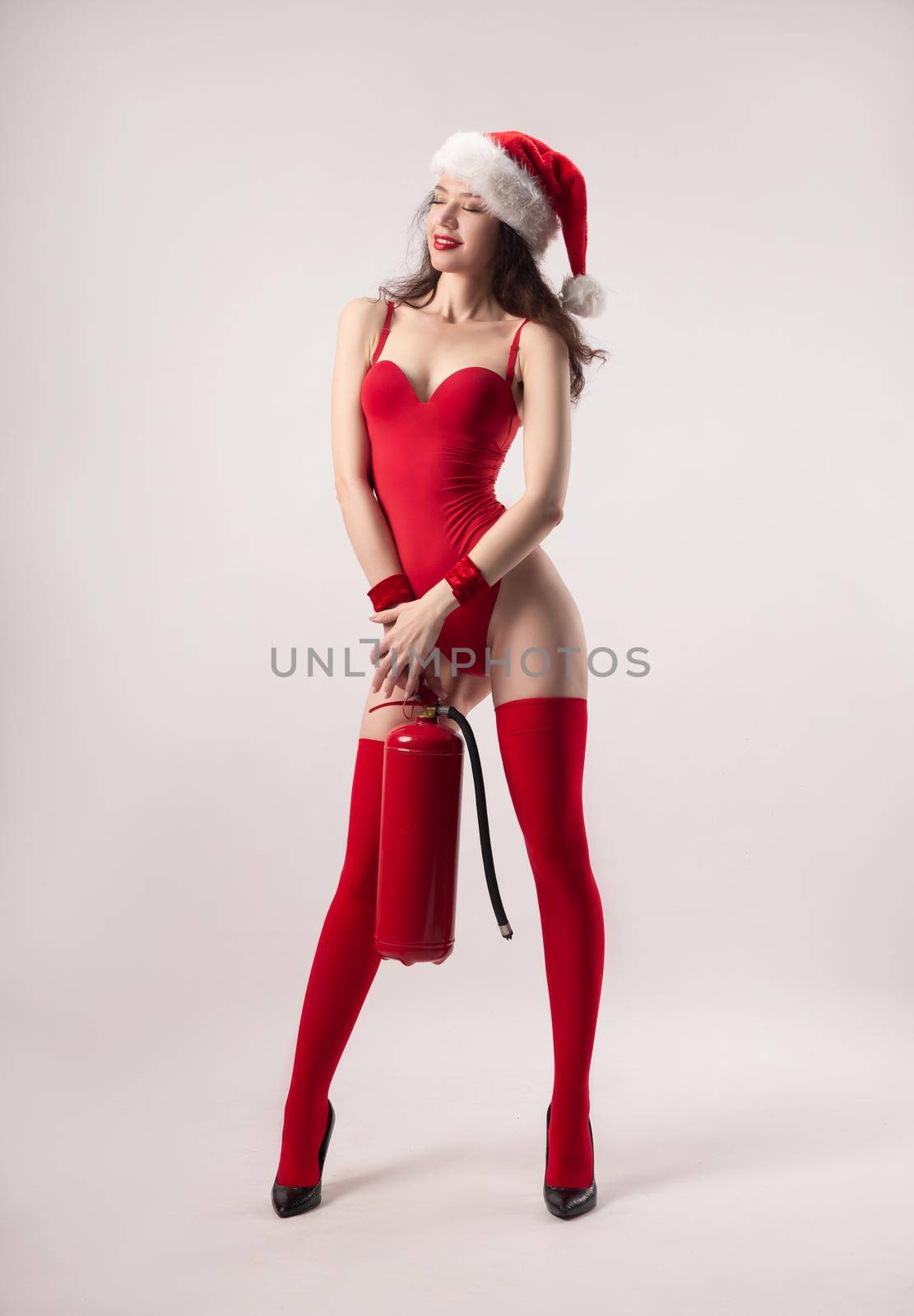 woman in a Santa Claus Christmas hat in a red bodysuit poses sexually with a red fire extinguisher