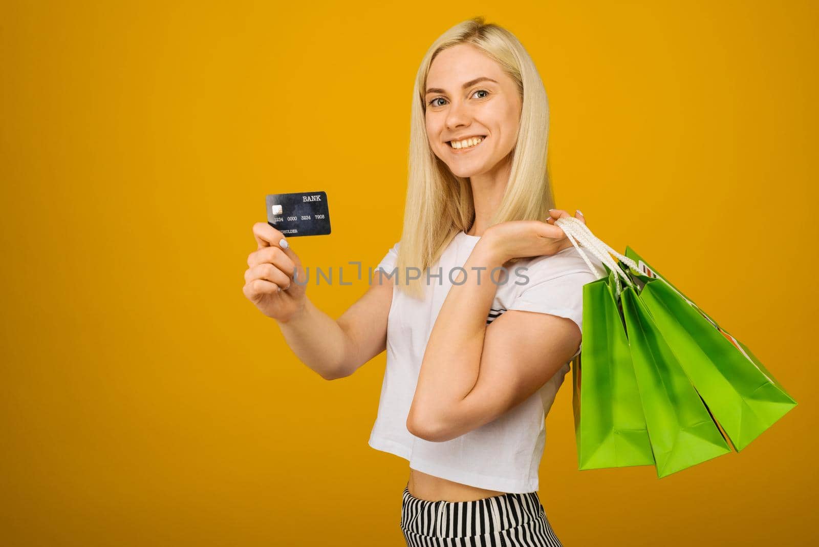 Close-up portrait of happy young beautiful blonde woman holding credit card and green shopping bags, looking at camera, isolated on yellow background - Image
