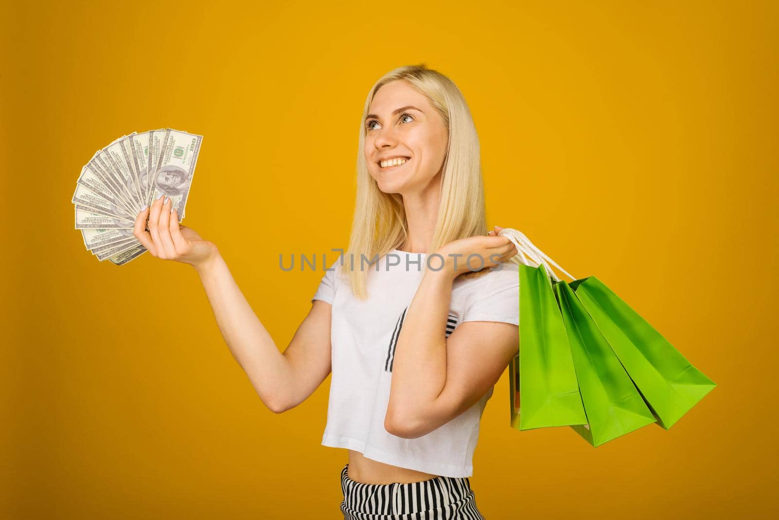 Close-up portrait of happy young beautiful blonde woman holding money and green shopping bags, isolated on yellow background - Image