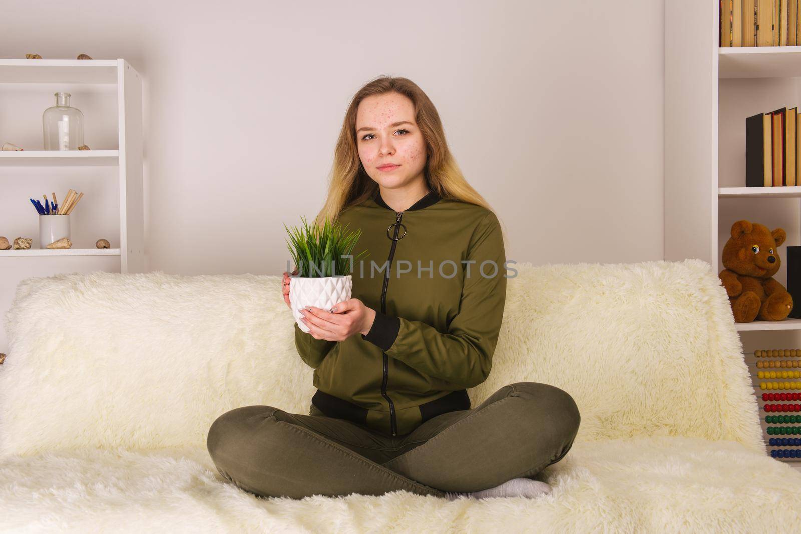 Young teen girl with acne face sitting on sofa and holding green houseplant - image