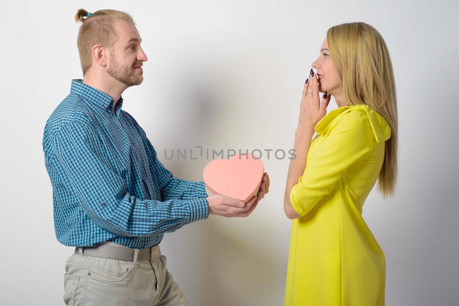 A stylish man in a checkered green shirt gives a beautiful slender girl in a yellow dress a gift box in the form of a heart. Happy couple in the studio.