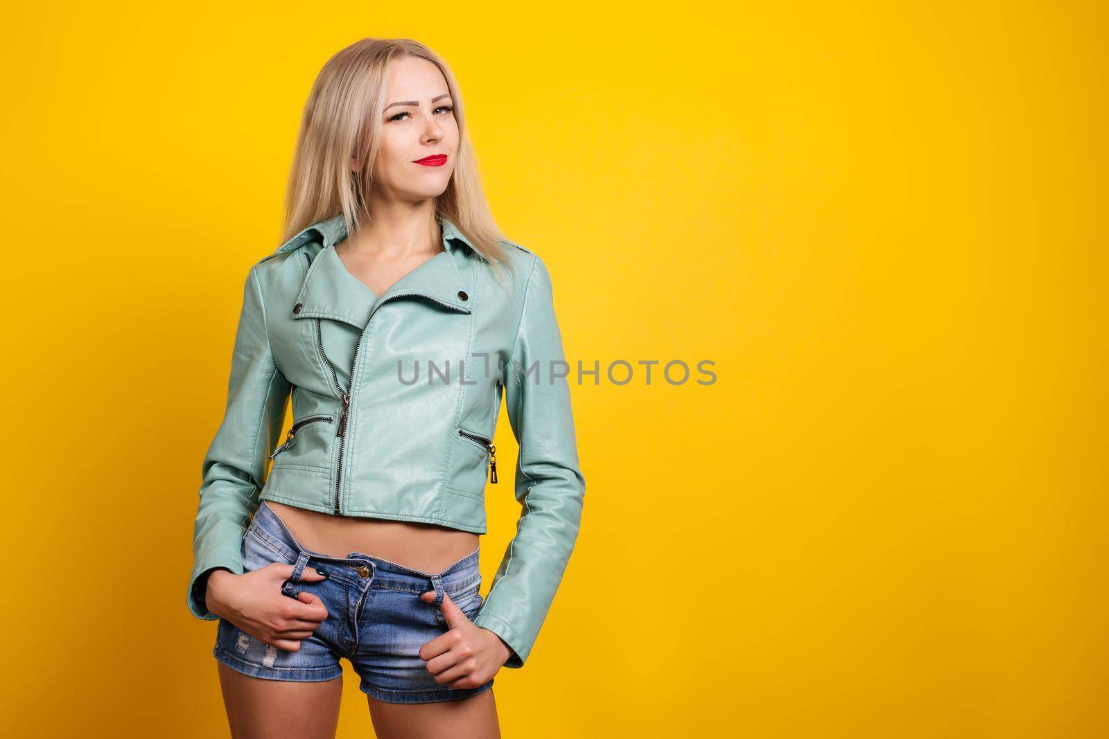 Sexy young woman in leather jacket and short denim shorts posing on a yellow background