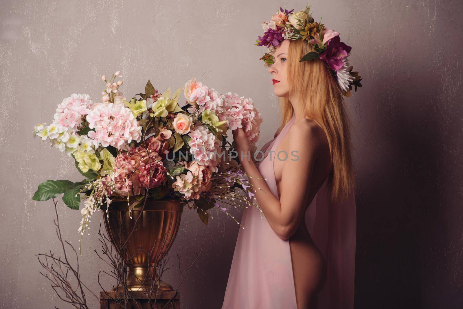 Beauty Summer model girl with colorful flowers wreath in elegant transparent pink fabric on the body stands next to a pot of flowers on a wooden box.