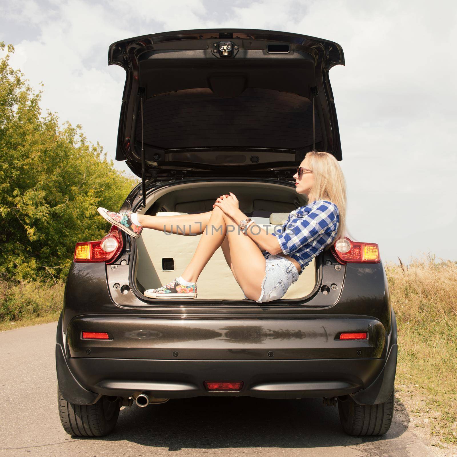 Young attractive woman sitting in the open trunk of a car. Summer road trip by zartarn
