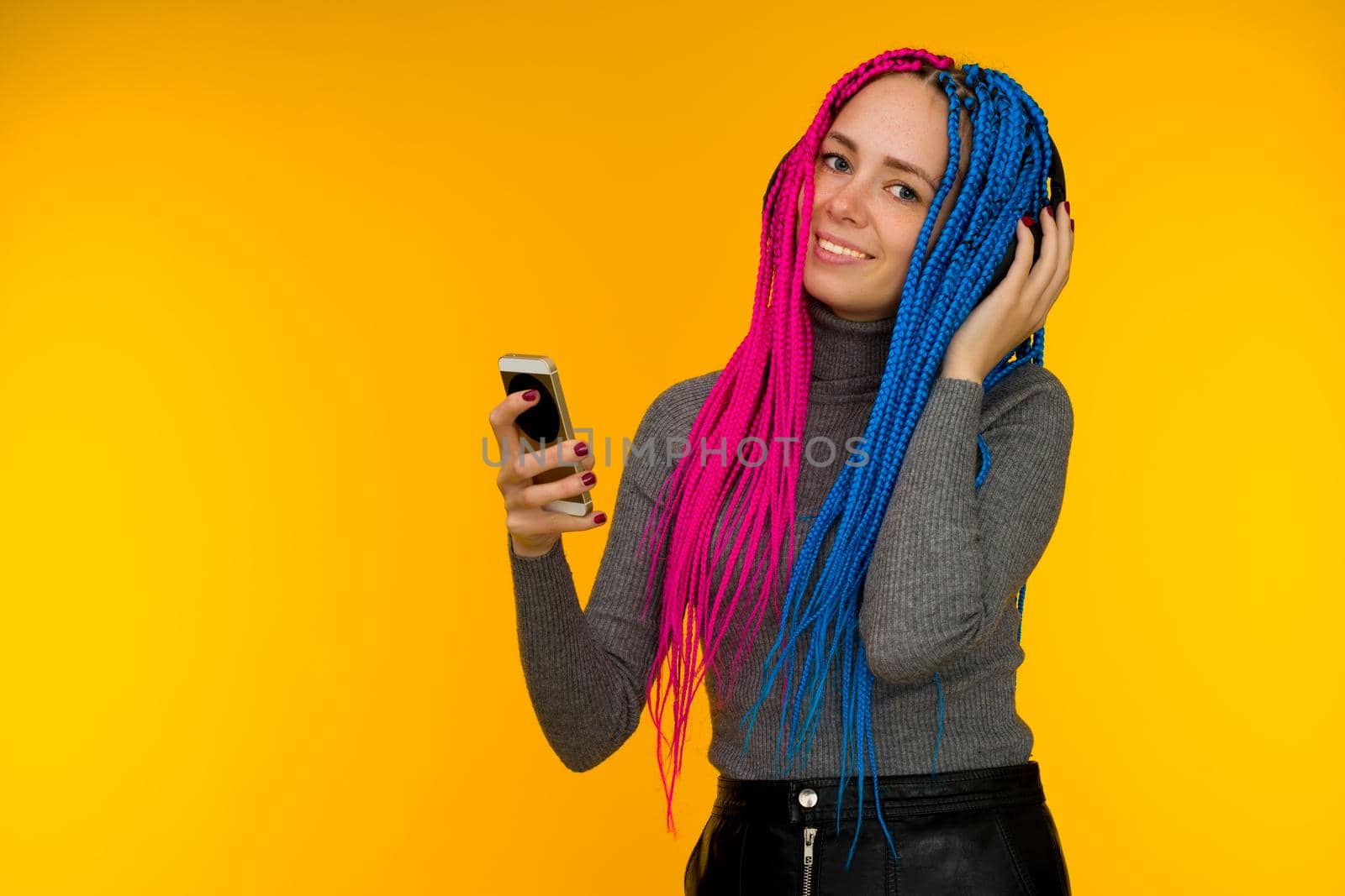 Happy cheerful woman with senegalese braids and freckles wearing wireless headphones listening to music from smartphone studio shot isolated on yellow background