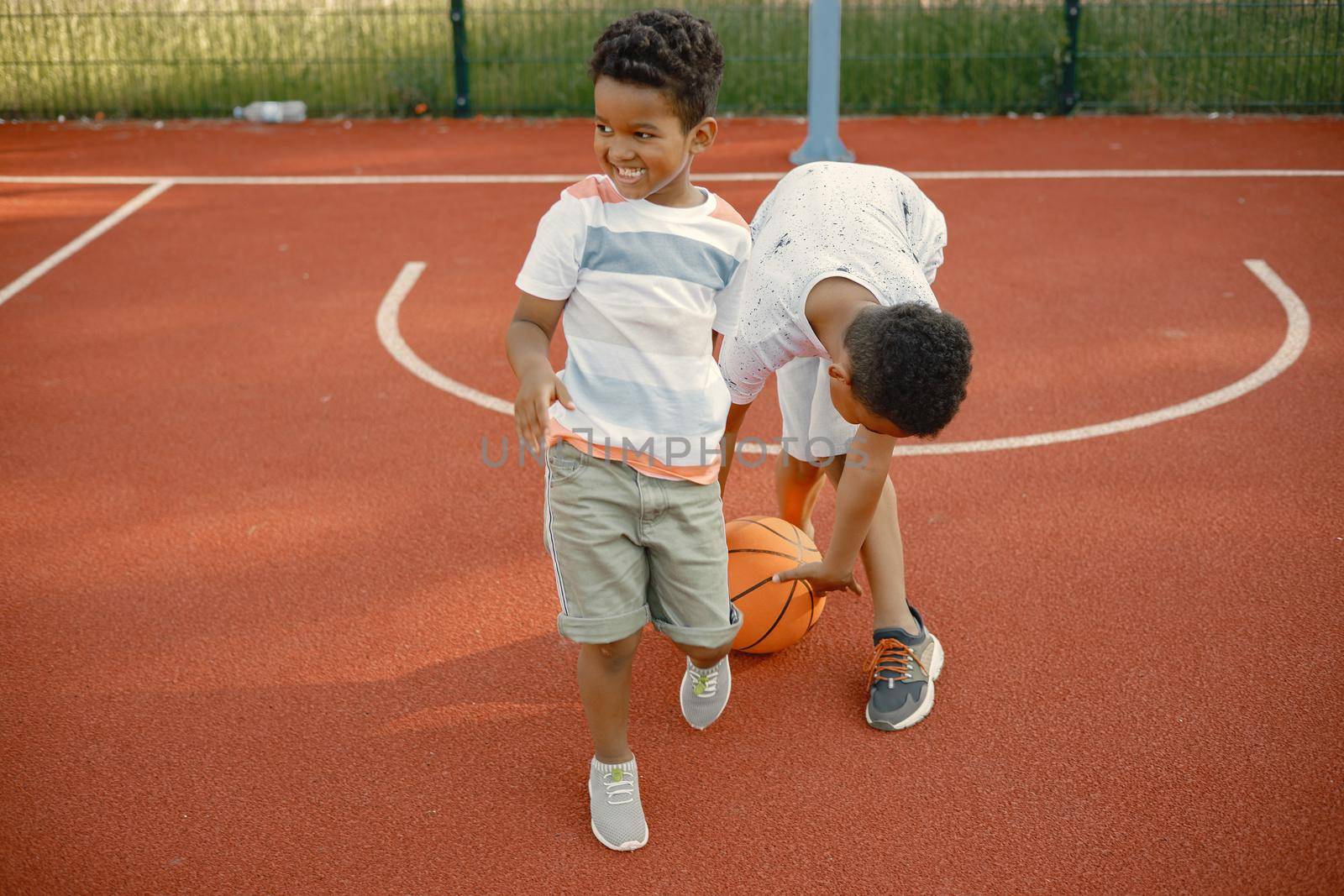 Two multiracional brothers playing basketball on a court near the park. Boys wearing white t-shirts. Older brother teach little one how to play basketball.