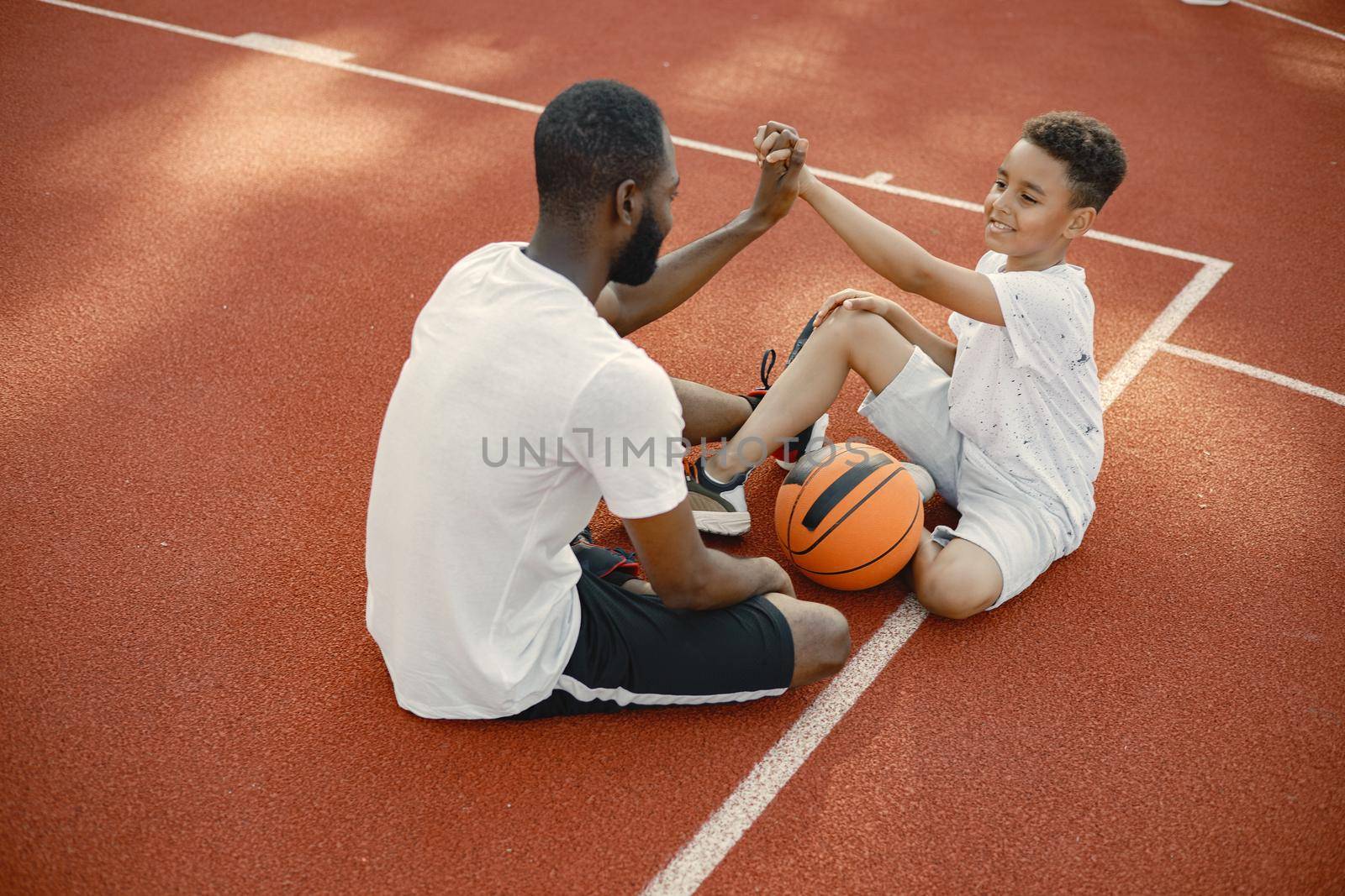 Young father and his son sitting on basketball court near the park. Man and boy wearing white t-shirts. They giving a high five to each other.