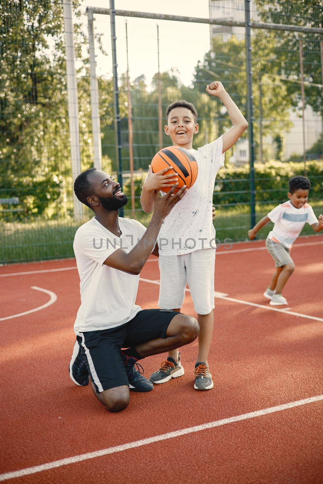Young father and his son standing on basketball court near the park. Boy posing for a photo pretending he is a winner. There are other boy behind.