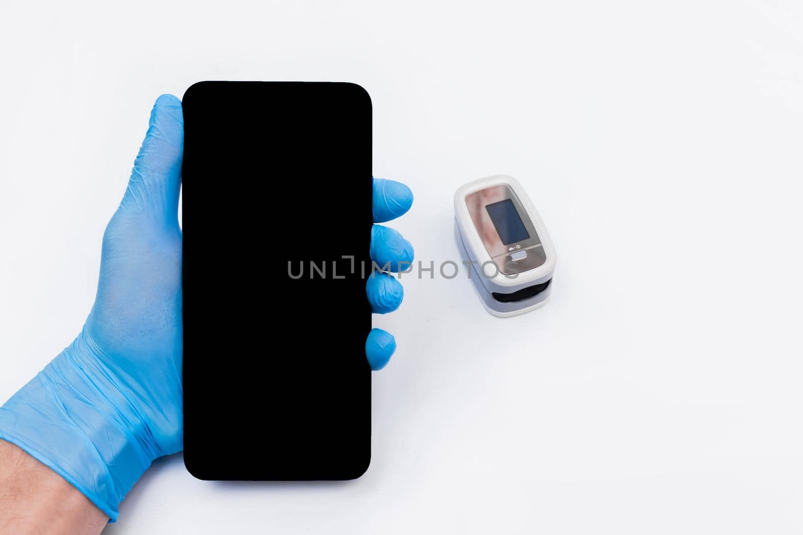 The hand of a medical doctor in a latex, rubber glove holds a mobile phone or smartphone with a screen against the background of the pulse oximeter.