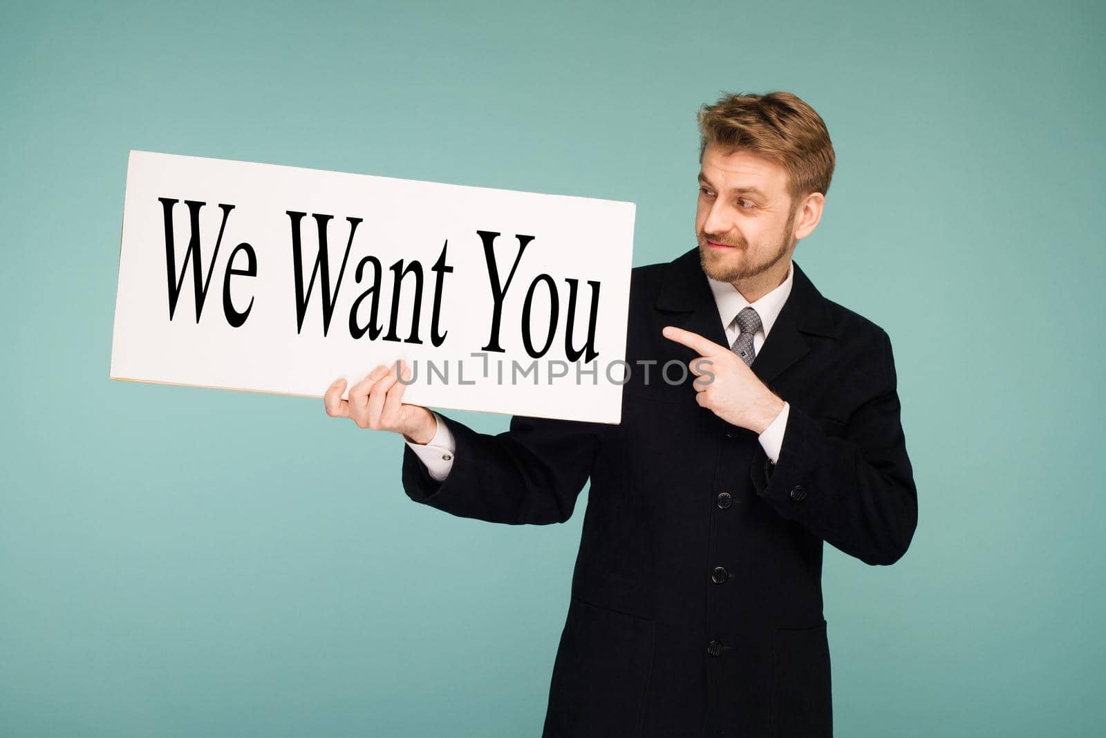 Happy smiling young business man points finger signboard with sign We Want You, on blue background