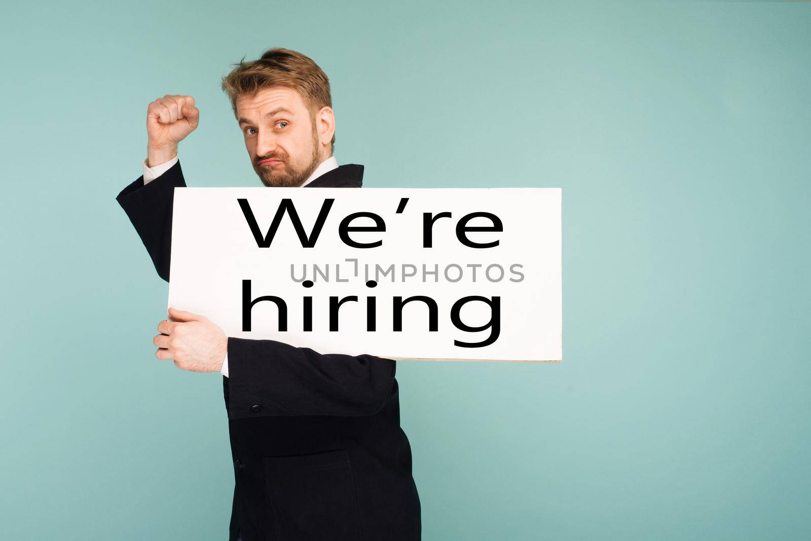 Funny young business man showing signboard with sign we hiring, on blue background