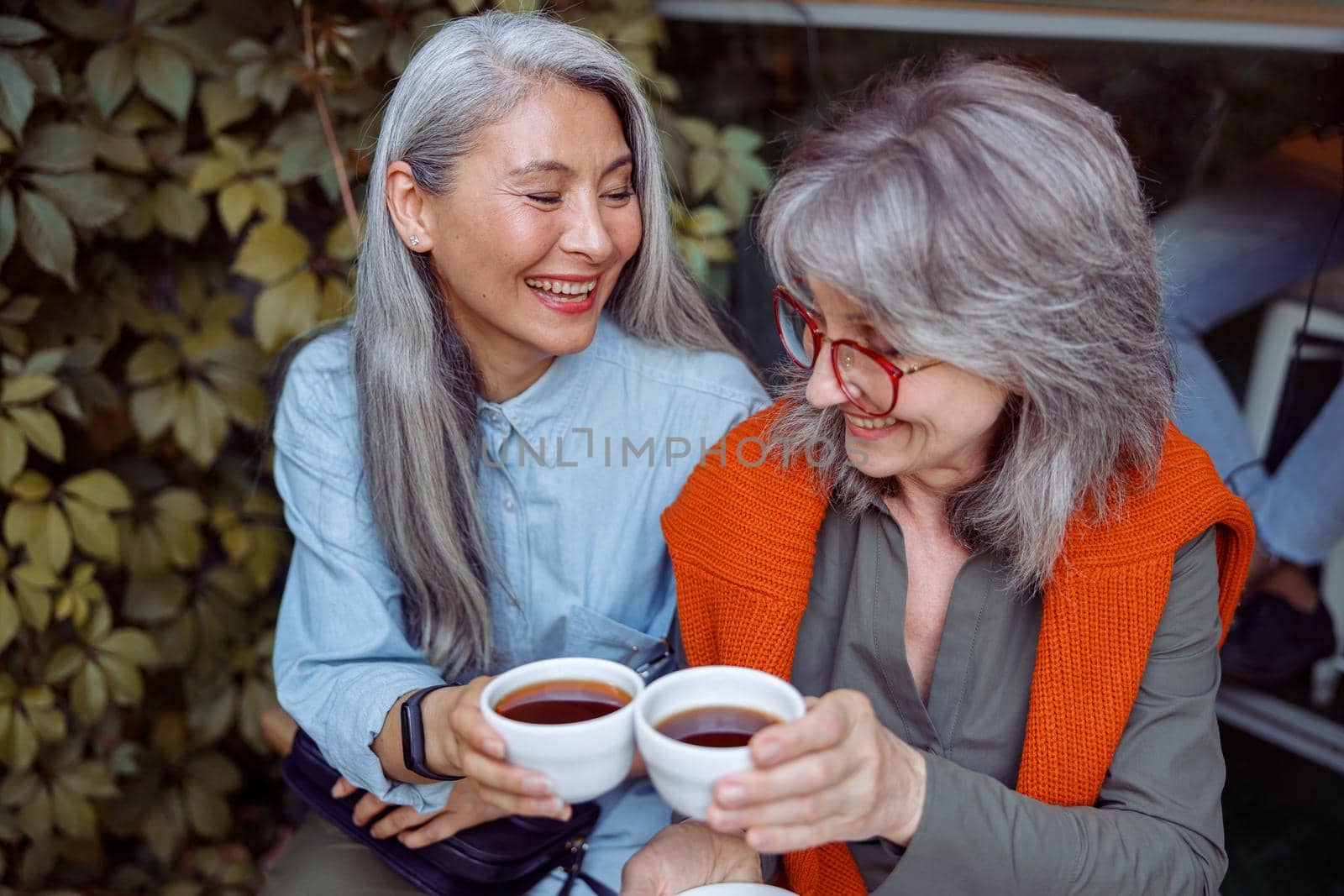 Happy senior Asian woman with grey haired friend clink cups of delicious beverages sitting on near cozy cafe outdoors. Long-time friendship