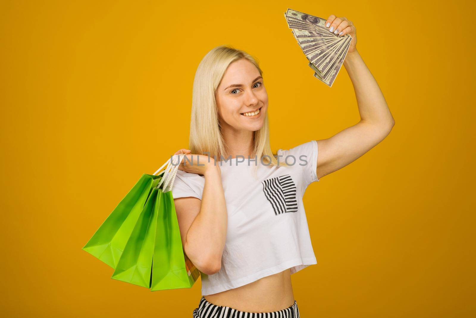 Close-up portrait of happy young beautiful blonde woman holding money and green shopping bags, looking at camera, isolated on yellow background - Image