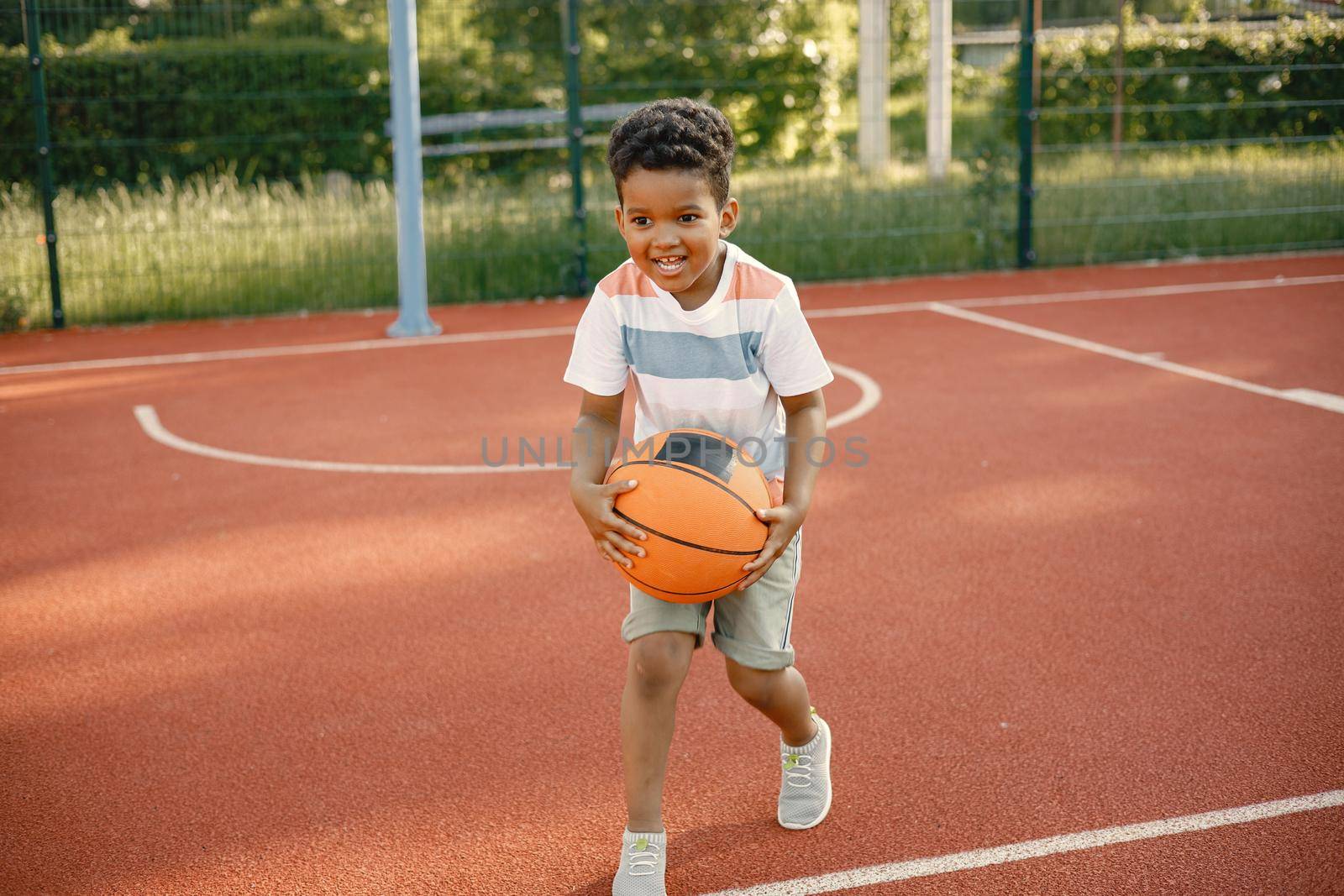 Young boy standing on basketball court near the park. Boy wearing white t-shirt. Boy playing with basketball ball.