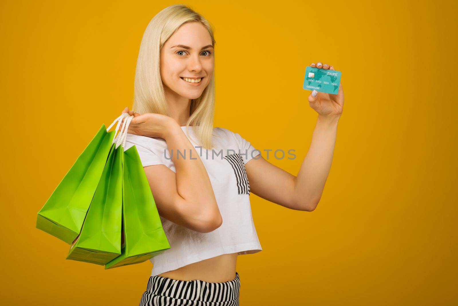 Close-up portrait of happy young beautiful blonde woman holding credit card and green shopping bags, looking at camera, isolated on yellow background by zartarn