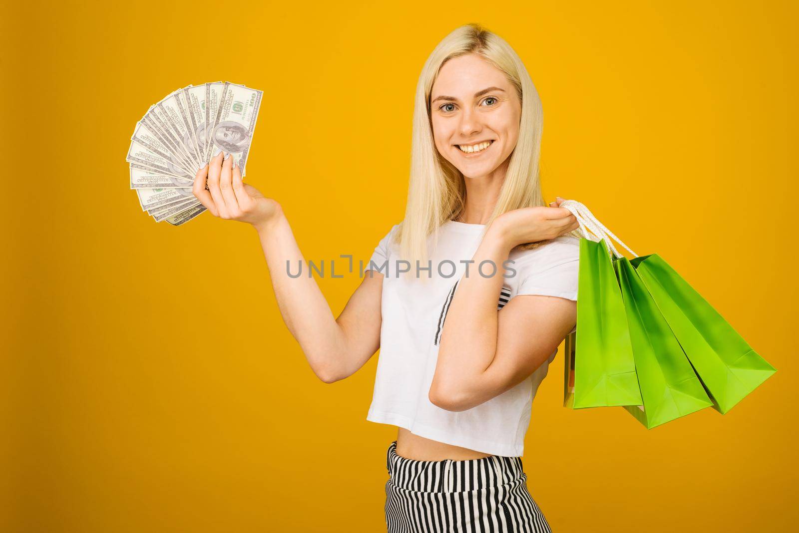 Close-up portrait of happy young beautiful blonde woman holding money and green shopping bags, looking at camera, isolated on yellow background - Image