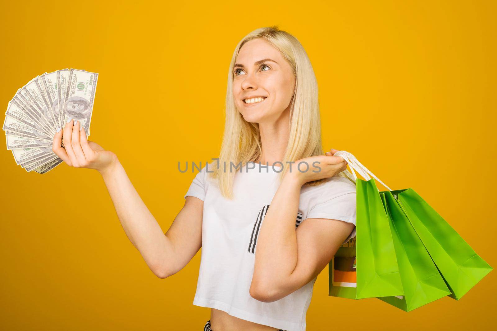 Close-up portrait of happy young beautiful blonde woman holding money and green shopping bags, isolated on yellow background - Image