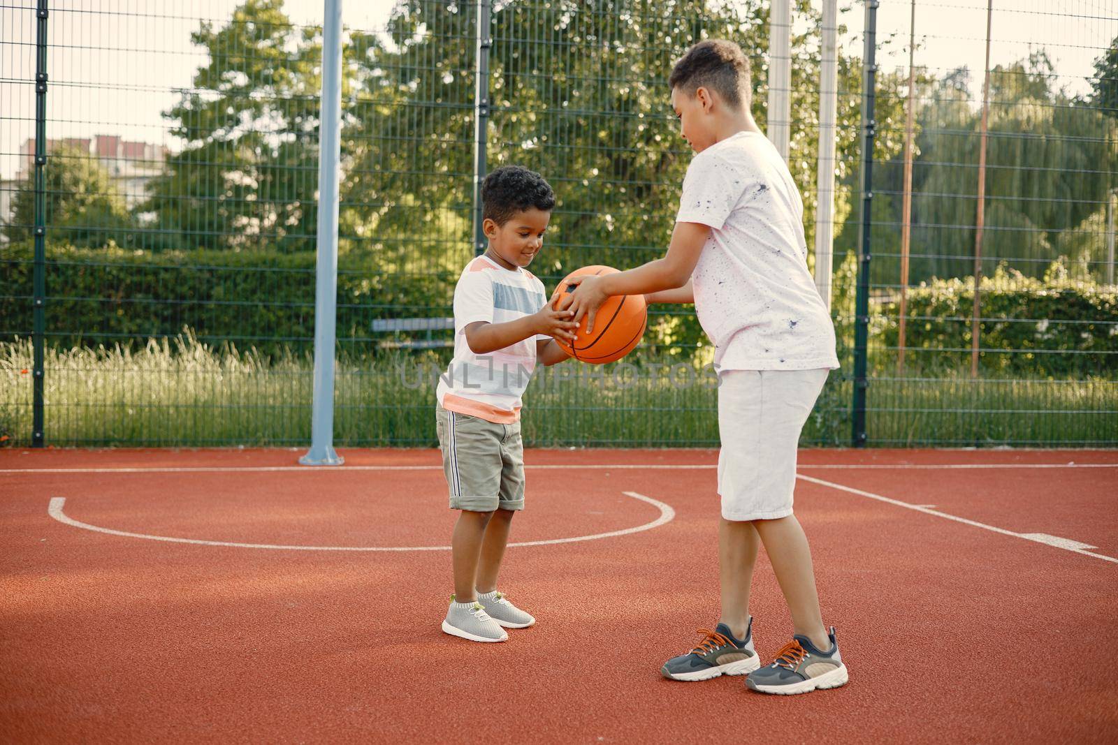 Two multiracional brothers playing basketball on a court near the park. Boys wearing white t-shirts. Older brother teach little one how to play basketball.