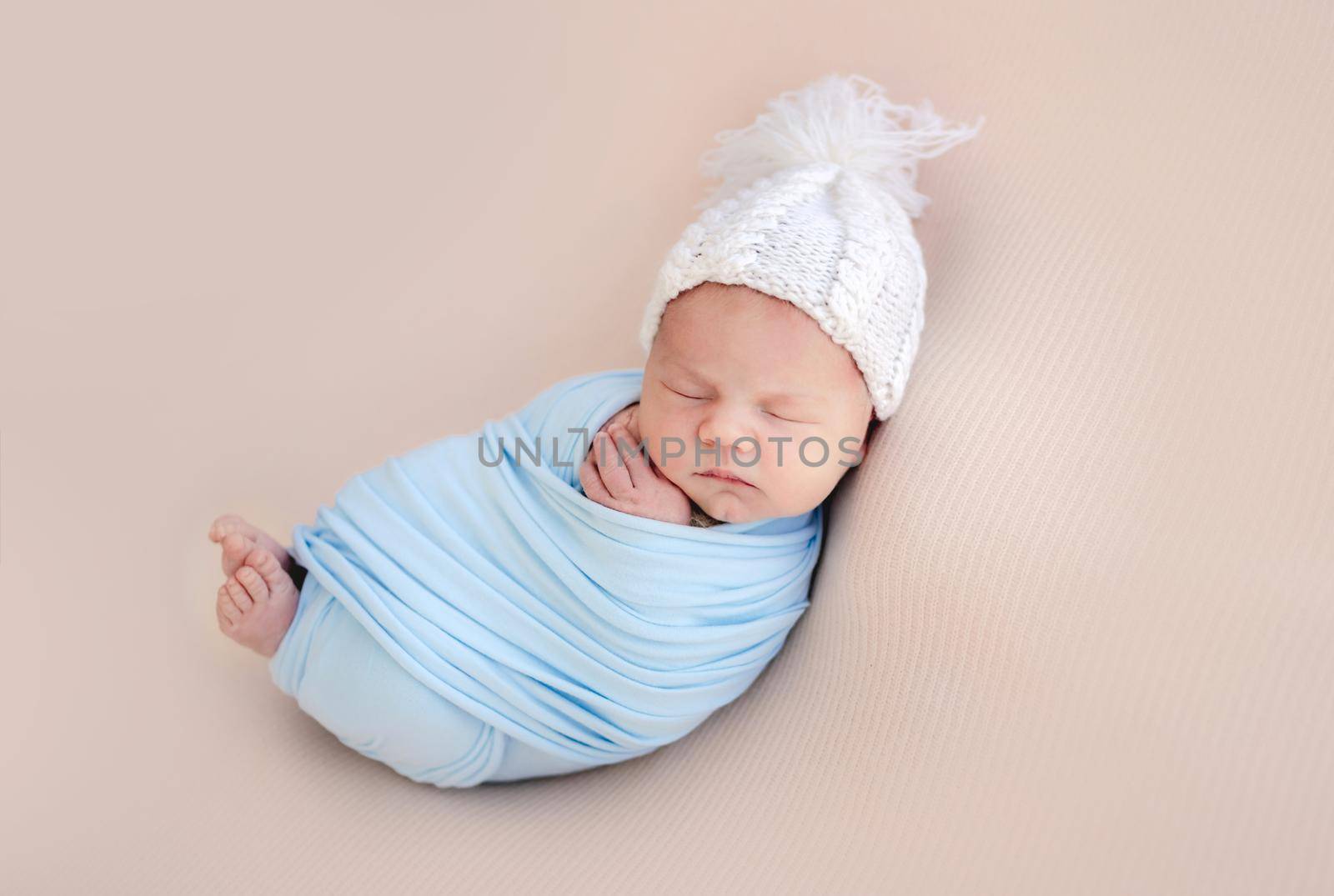 Newborn baby boy swaddled in blue fabric and wearing hat sleeping. Infant child kid napping during studio photoshoot