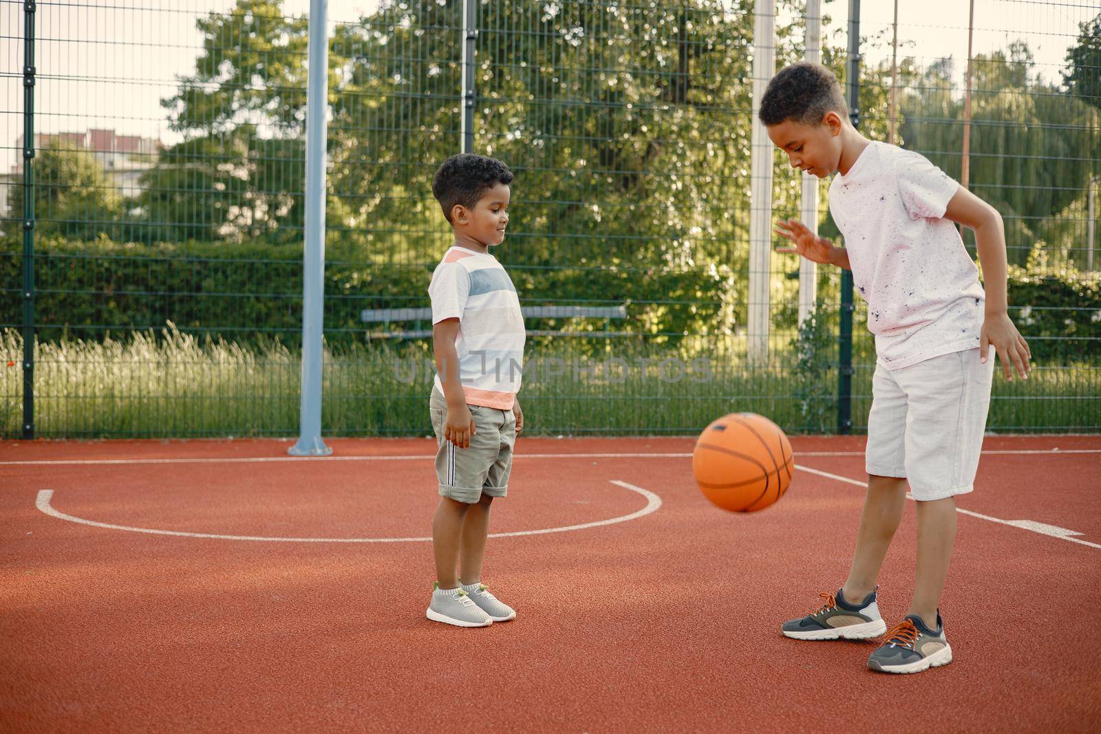 Two multiracional brothers playing basketball on a court near the park. Boys wearing white t-shirts. Older brother teach little one how to play basketball.
