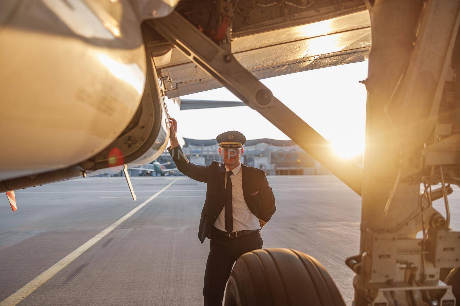 Professional male pilot in uniform and hat looking at camera, leaning on a big passenger airplane ready for departure in airport at sunset. Aircraft, occupation, transportation concept